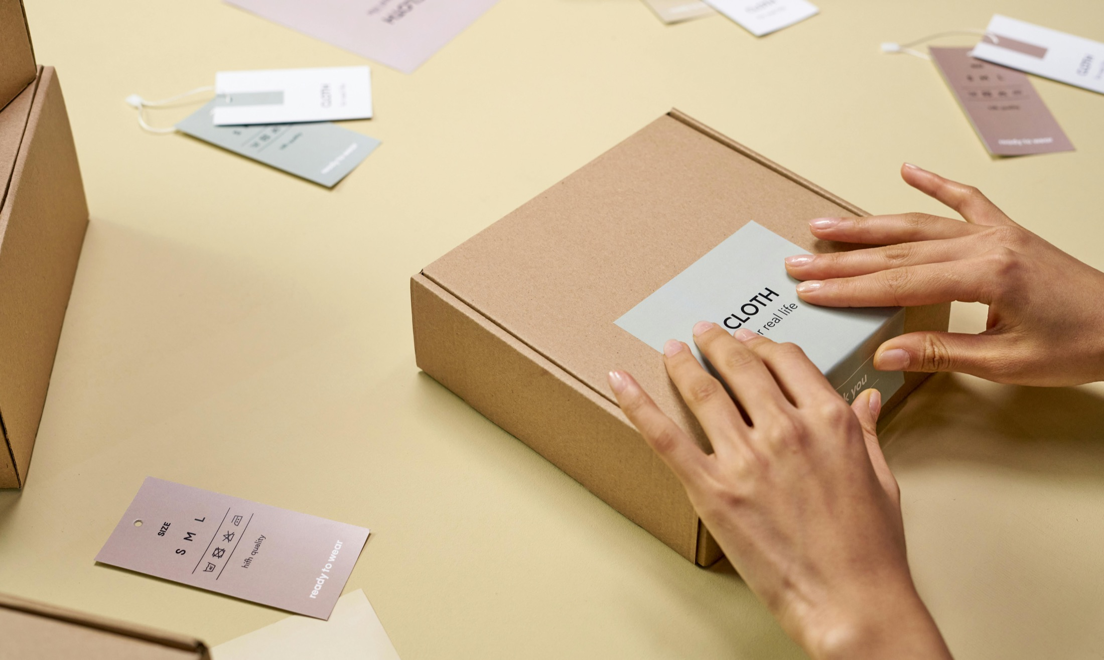 Hands applying a label to a cardboard product box with hang tags and packaging samples nearby